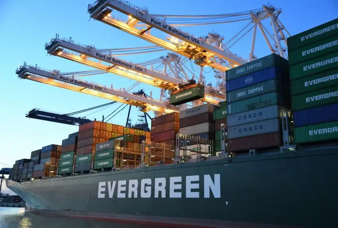 Statek towarowy „Evergreen” w porcie w Baltimore w Stanach Zjednoczonych. Liczne kontenery ułożone w stosy pod dużymi dźwigami na tle czystego wieczornego nieba.